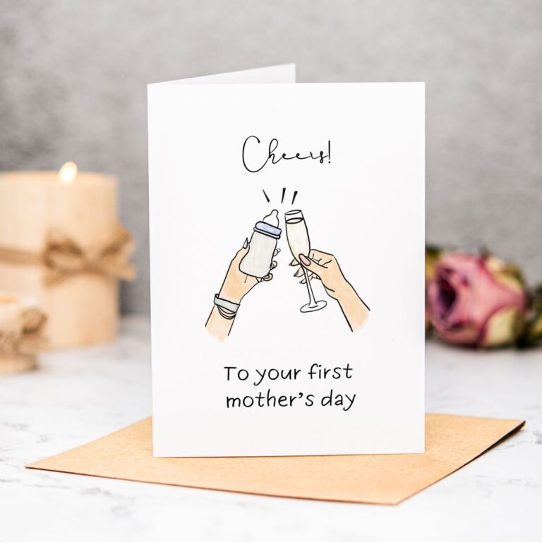 A First Mothers Day Card featuring two hands toasting—one with a baby bottle, the other with a champagne glass. The card reads Cheers! To your first mother’s day, with a blurred candle and flower in the background.