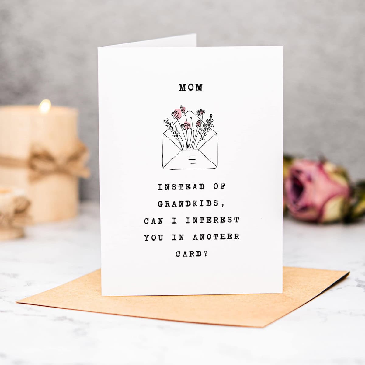 A Free Funny Mothers Day Card on a brown envelope reads, “MOM. Instead of grandkids, can I interest you in another card?” with a flower-filled envelope illustration. A lit candle and rose decorate the background.
