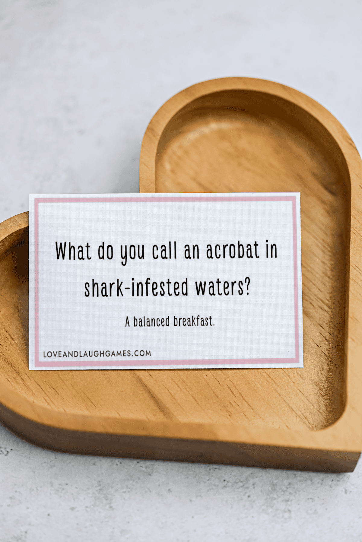 A card on a wooden heart-shaped tray reads: What do you call an acrobat in shark-infested waters? A balanced breakfast. Perfect for fans of Acrobat Jokes!.