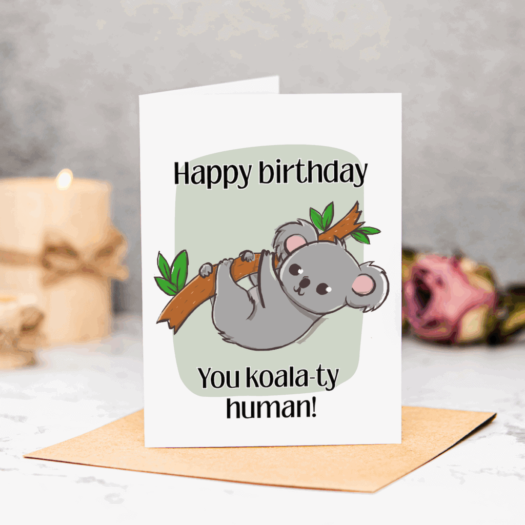 A greeting card with a cartoon koala hanging from a branch, captioned “Happy birthday. You koala-ty human!” Perfect as a New Home Printable Card, it stands on a brown envelope, with a candle and pink rose blurred in the background.