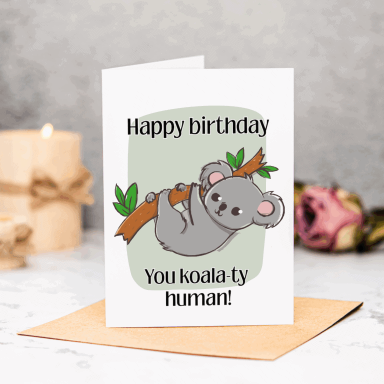 A greeting card with a cartoon koala hanging from a branch, captioned “Happy birthday. You koala-ty human!” Perfect as a New Home Printable Card, it stands on a brown envelope, with a candle and pink rose blurred in the background.
