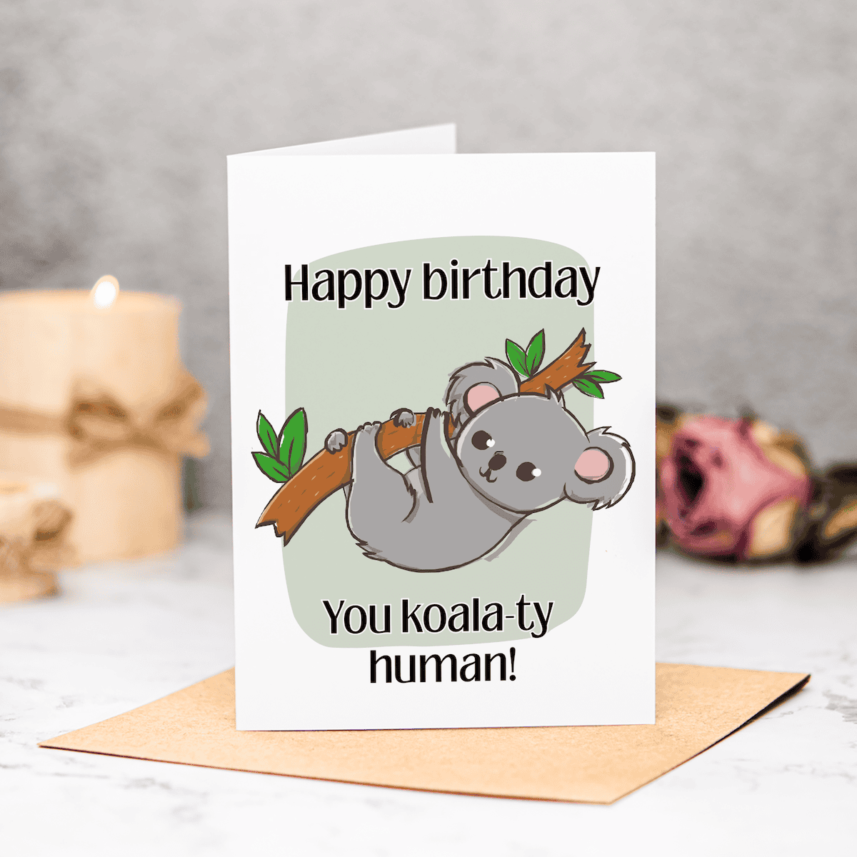 A greeting card with a cartoon koala hanging from a branch, captioned “Happy birthday. You koala-ty human!” Perfect as a New Home Printable Card, it stands on a brown envelope, with a candle and pink rose blurred in the background.