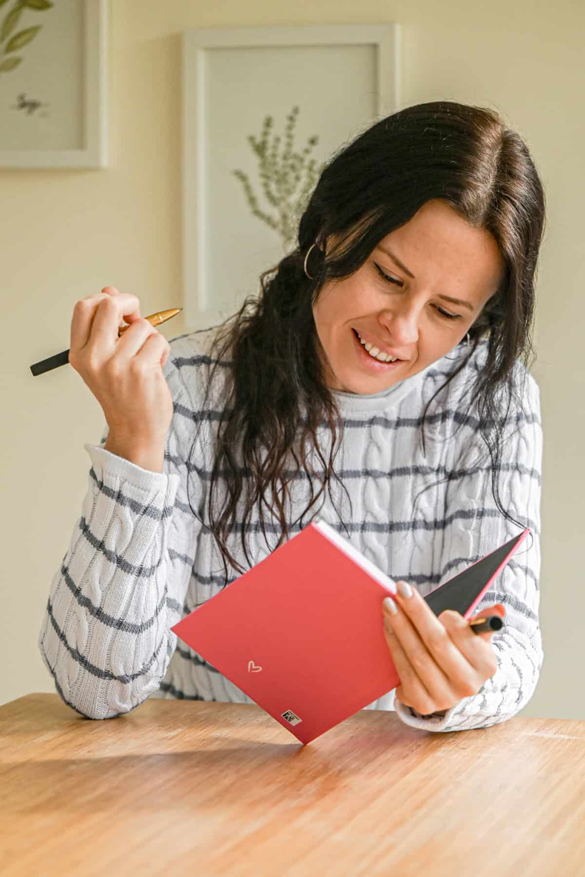A woman with long dark hair, wearing a white and gray striped sweater, smiles while writing in a red notebook at a wooden table. She holds a pen and looks at the open notebook. Framed art hangs on the wall behind her.
