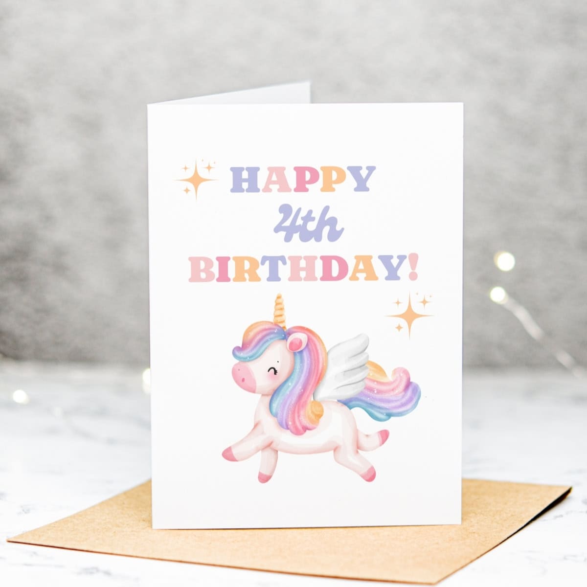 A whimsical Unicorn 4th Birthday Card featuring a flying unicorn with pastel rainbow wings. "Happy 4th Birthday!" appears in playful, colorful letters, surrounded by gold sparkles. The card is displayed on a brown envelope.
