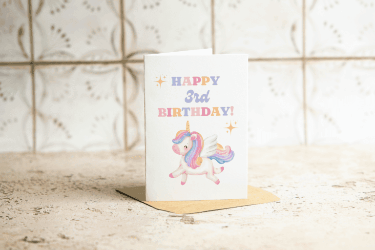 A birthday card standing on a brown envelope reads HAPPY 3rd BIRTHDAY! in colorful letters, with an illustration of a smiling unicorn with wings on the front. The background is a light, patterned surface.