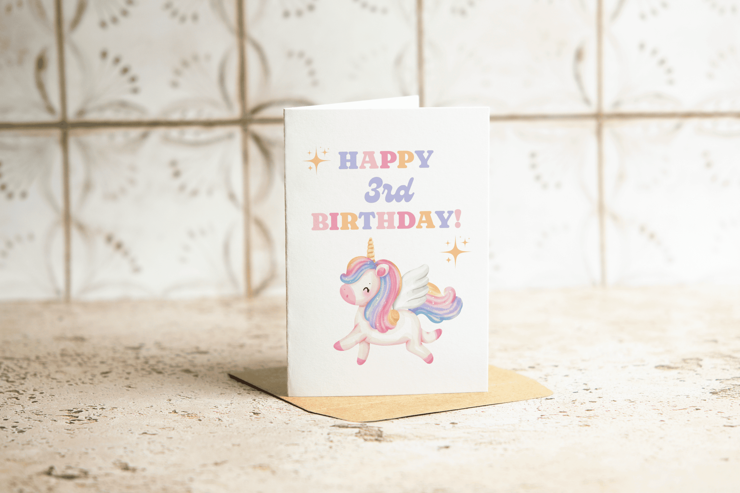 A birthday card standing on a brown envelope reads HAPPY 3rd BIRTHDAY! in colorful letters, with an illustration of a smiling unicorn with wings on the front. The background is a light, patterned surface.