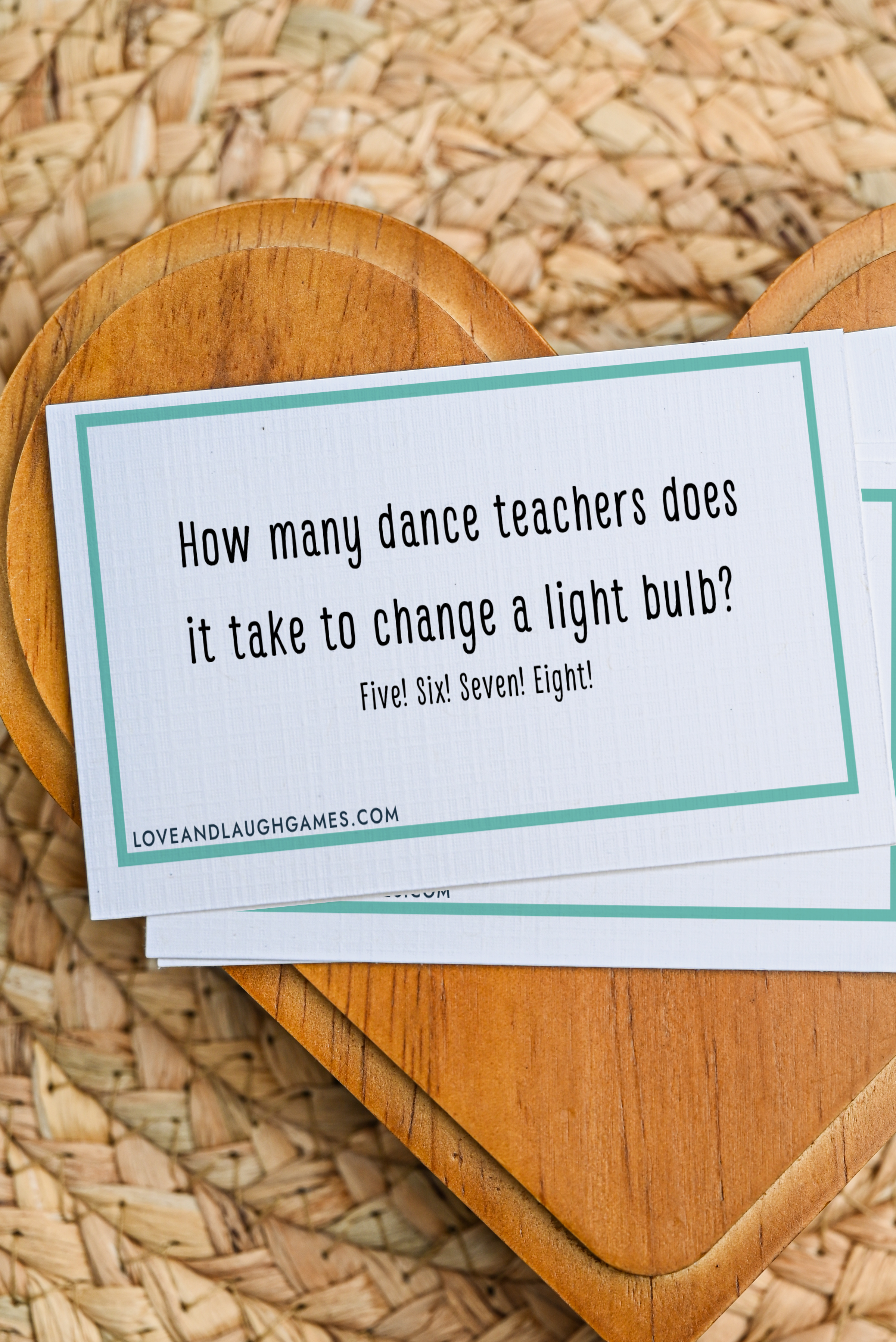 A card on a wooden heart reads: How many dance teachers does it take to change a light bulb? Five! Six! Seven! Eight! The card rests on a woven surface.