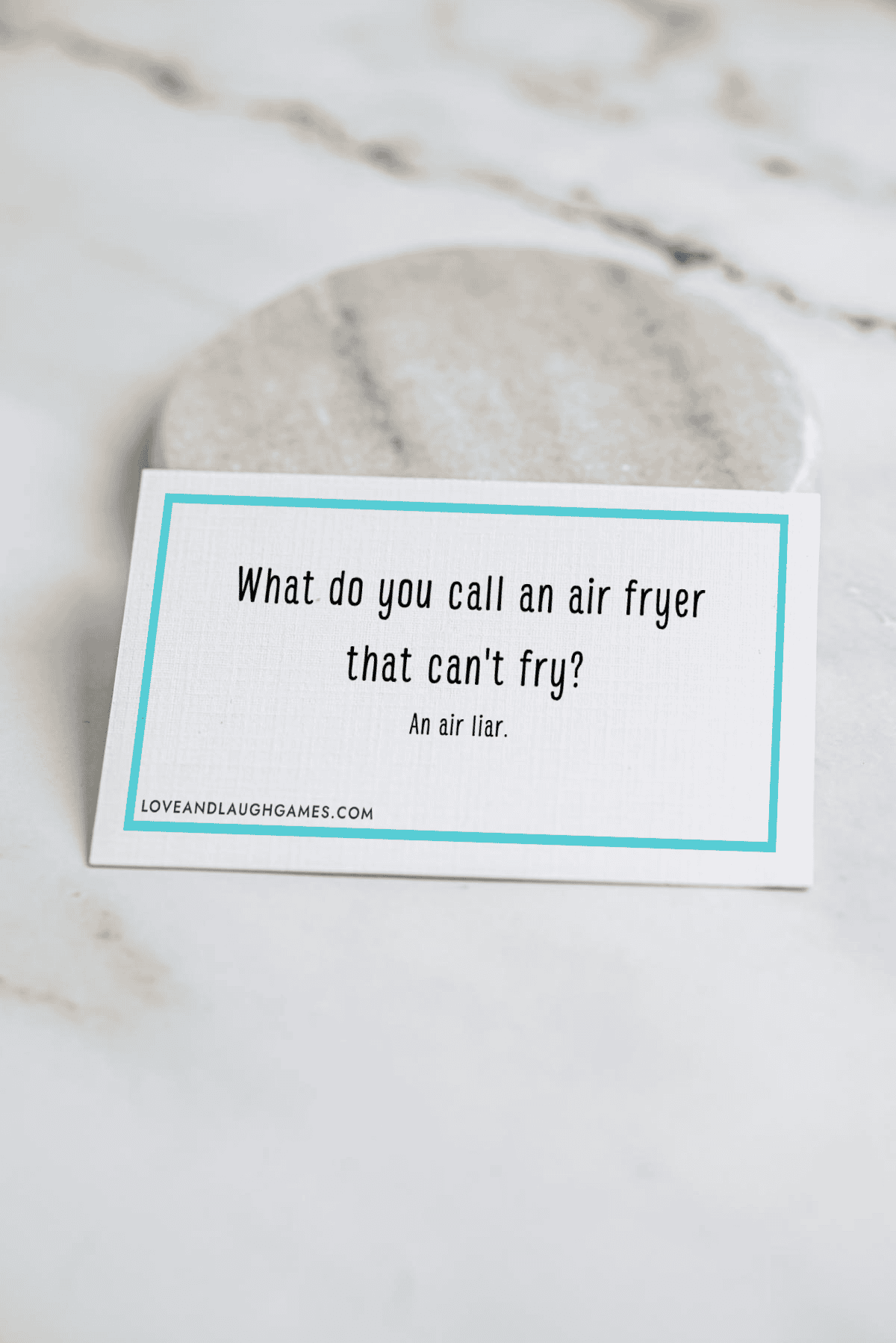 A card on a marble surface reads: What do you call an air fryer that cant fry? An air liar. The card has a blue border and the website loveandlaughgames.com at the bottom.