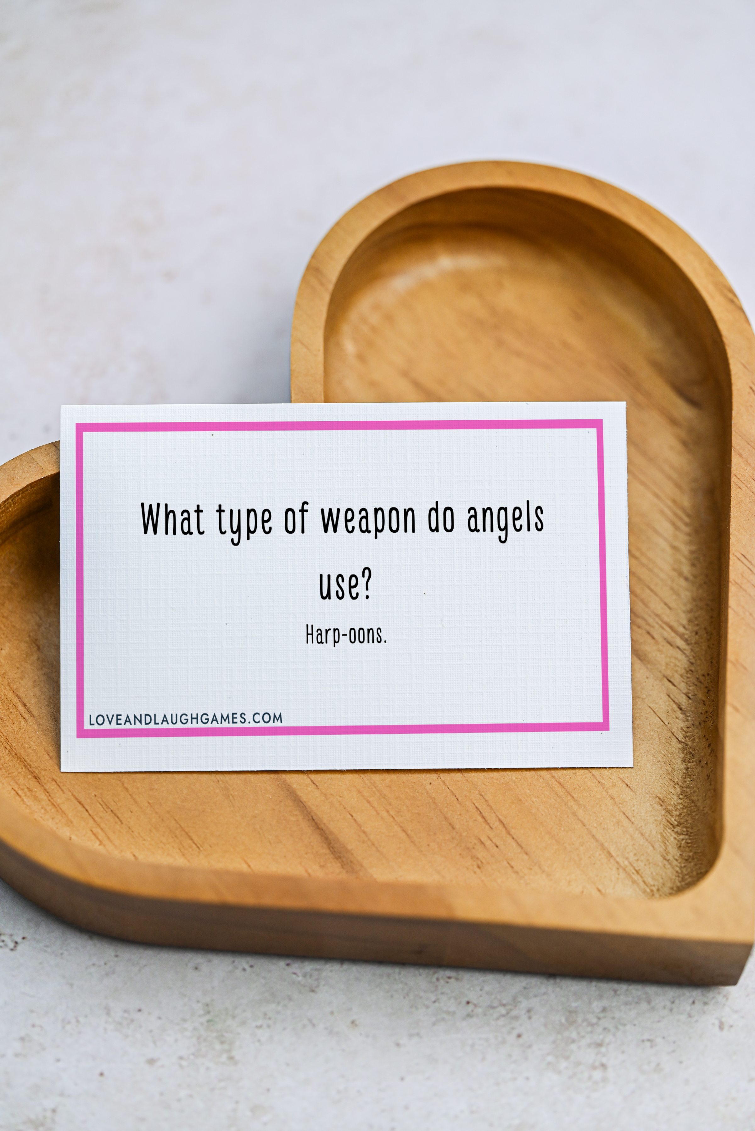A card with a pink border sits on a wooden heart-shaped tray. The card reads: What type of weapon do angels use? Harp-oons.
