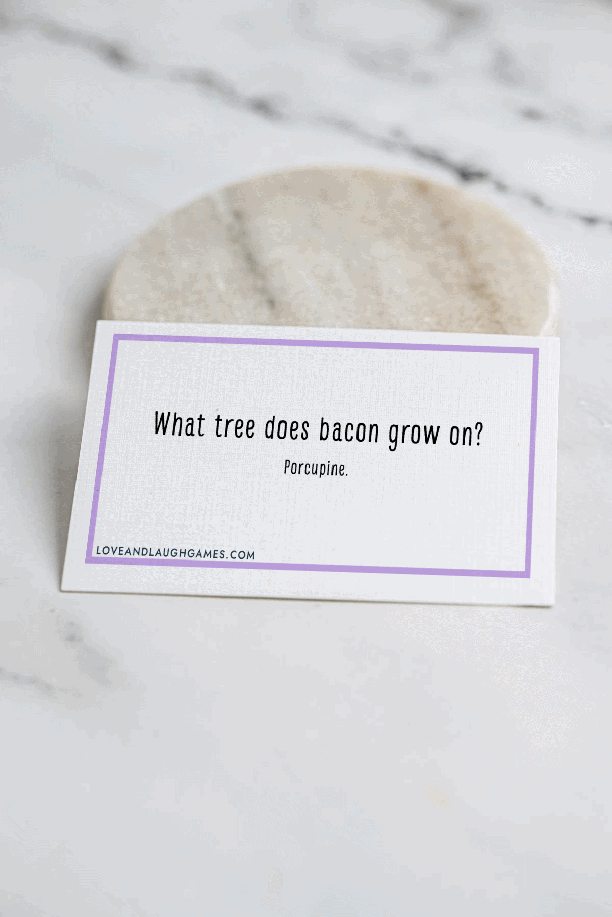 A card on a marble surface reads, What tree does bacon grow on? Porcupine. The card has a purple border and a website link at the bottom.