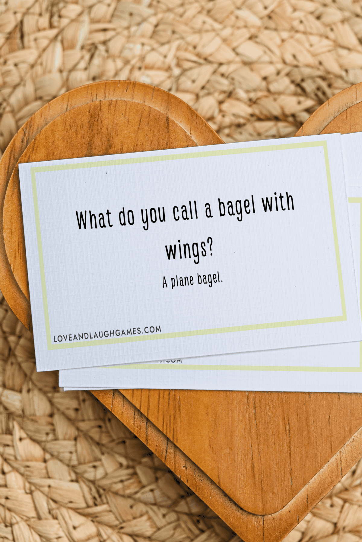 A card with the text: “What do you call a bagel with wings? A plane bagel.” placed on a wooden heart-shaped board, with a woven surface underneath.