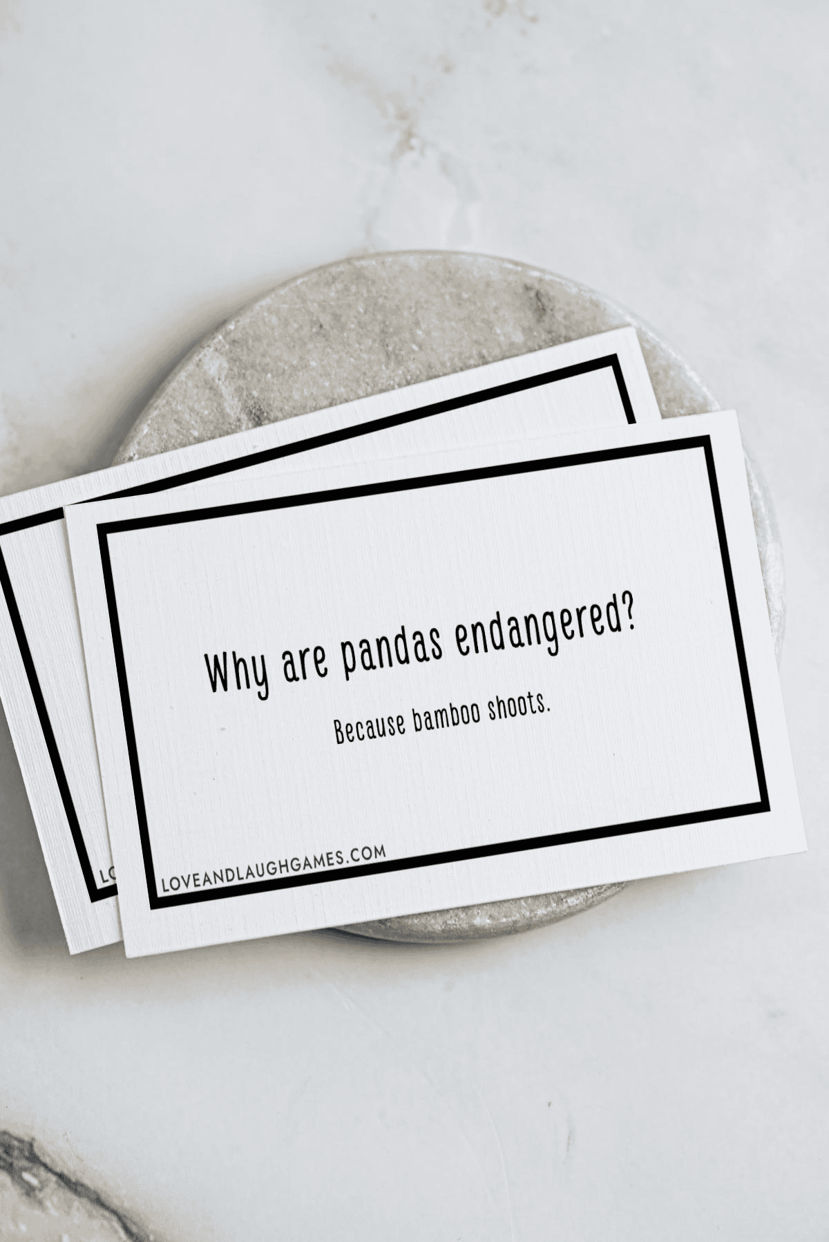 Two white cards with black borders are stacked on a round stone surface. The top card reads, Why are pandas endangered? Because bamboo shoots. The website loveandlaughgames.com is printed at the bottom.