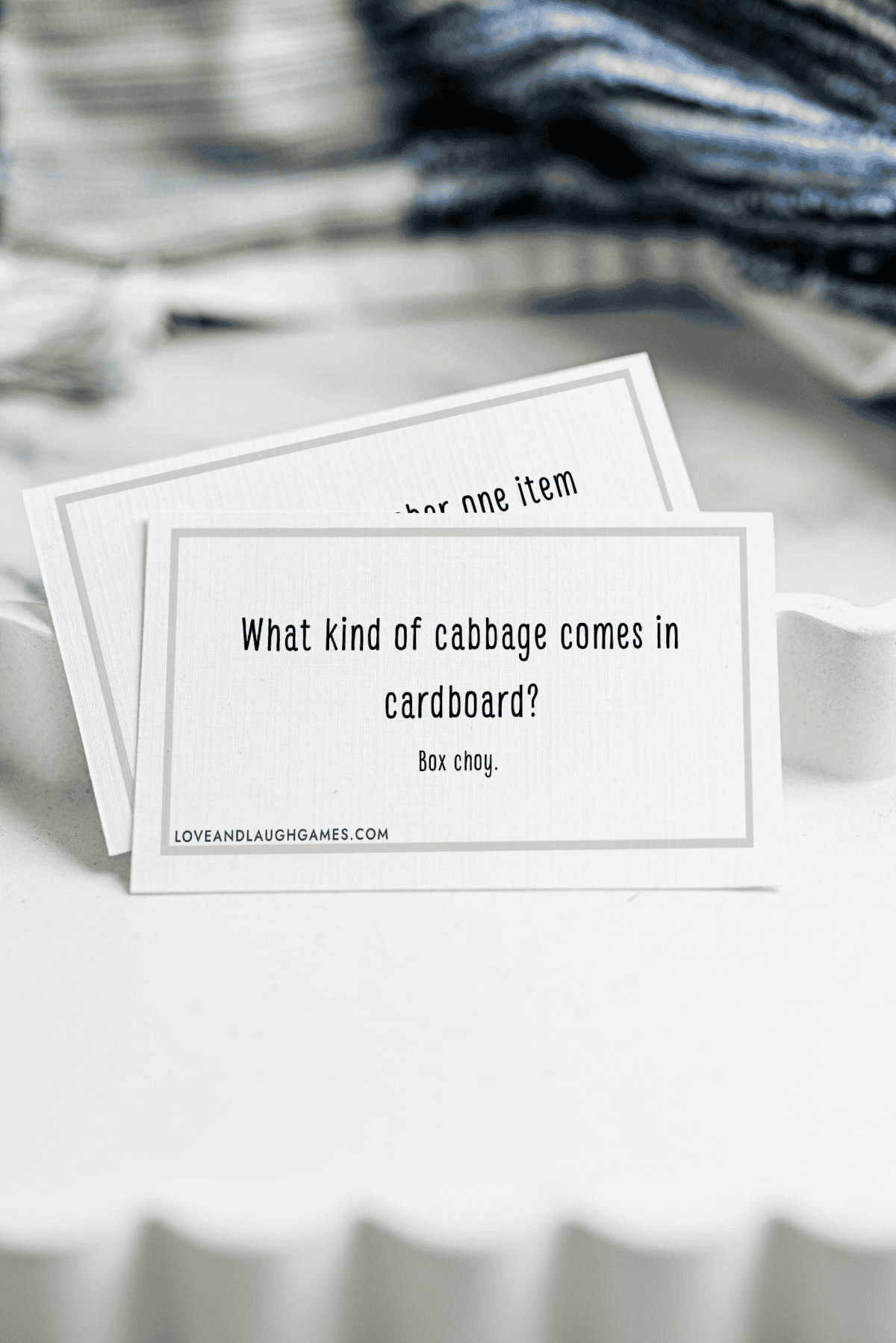 A card with the joke What kind of cabbage comes in cardboard? Box choy. sits in a white dish with another card partially visible behind it; blue and white fabric is in the background.
