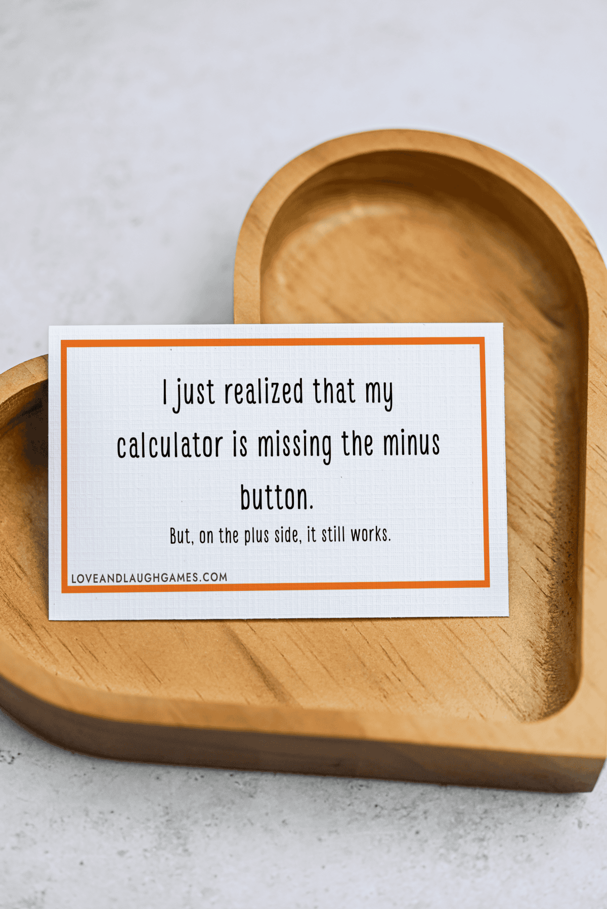 A card with a joke: I just realized that my calculator is missing the minus button. But, on the plus side, it still works. The card rests on a wooden dish shaped like a heart.
