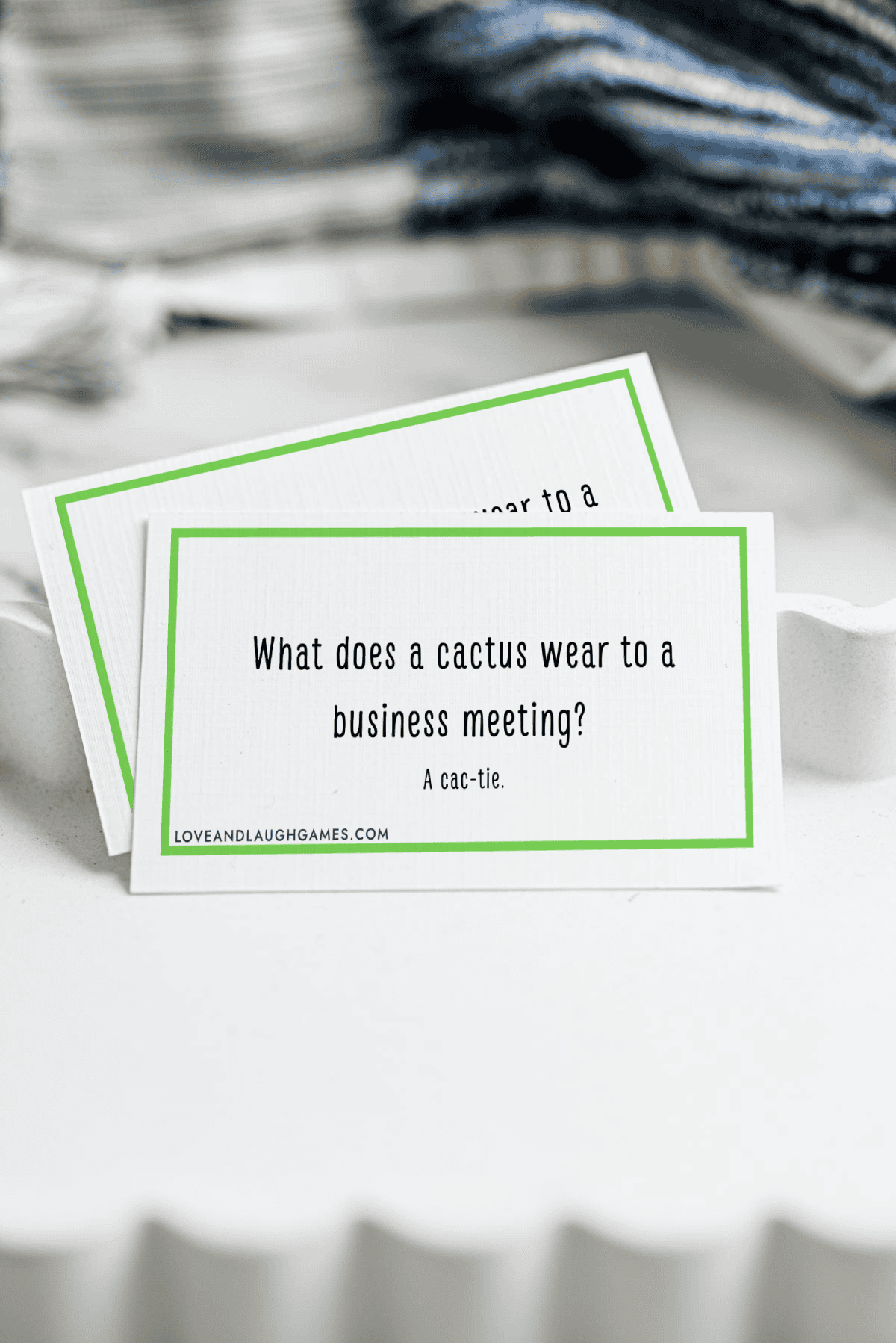 A card with the joke, What does a cactus wear to a business meeting? A cac-tie. is displayed on a white wavy dish, with another card and folded fabric visible in the background.