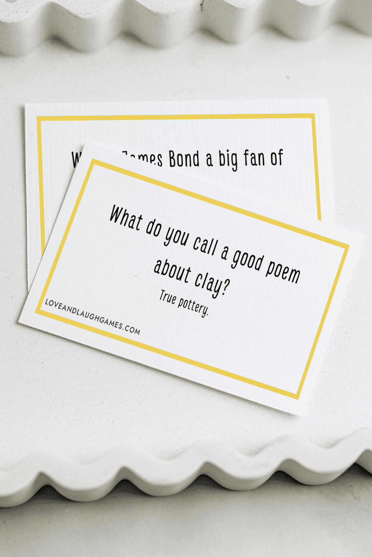 Two white cards with yellow borders are shown. The card on top reads, What do you call a good poem about clay? True pottery. The second card is partially visible beneath it.
