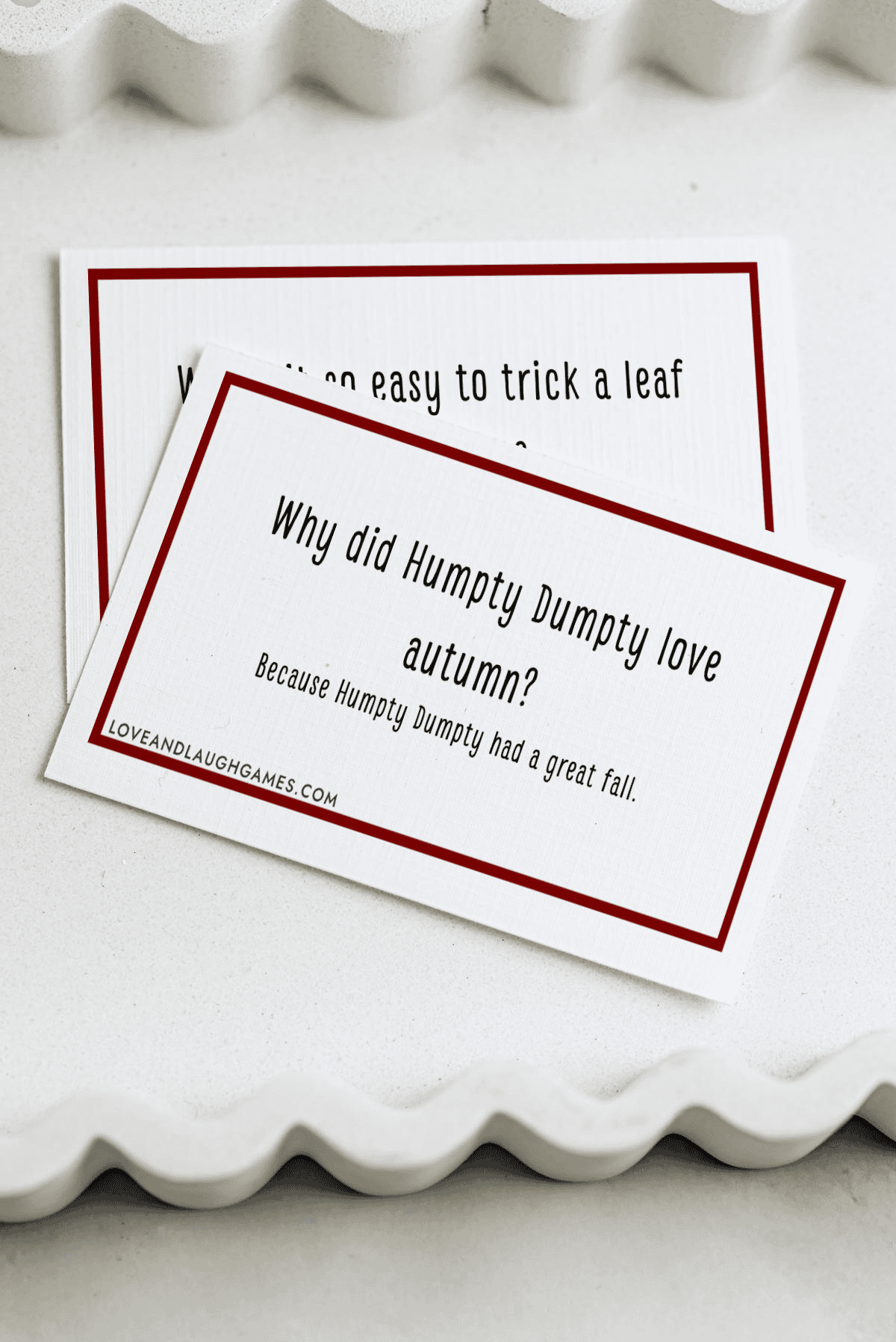 A card with the joke: Why did Humpty Dumpty love autumn? Because Humpty Dumpty had a great fall. Another partially hidden card is underneath, all on a white wavy-edged tray.