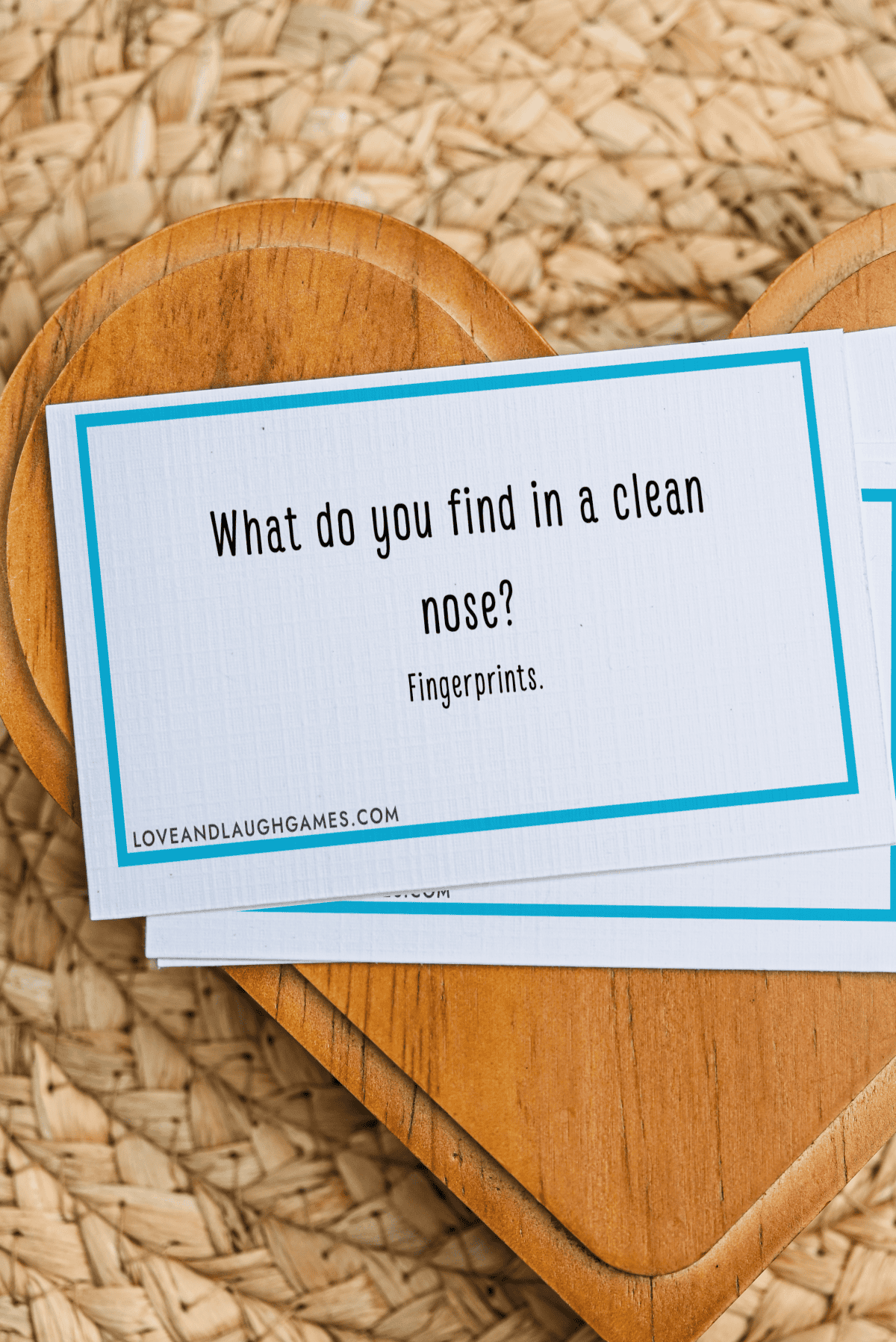 A card on a wooden heart reads, What do you find in a clean nose? Fingerprints. The card is white with a blue border and sits on a woven surface.
