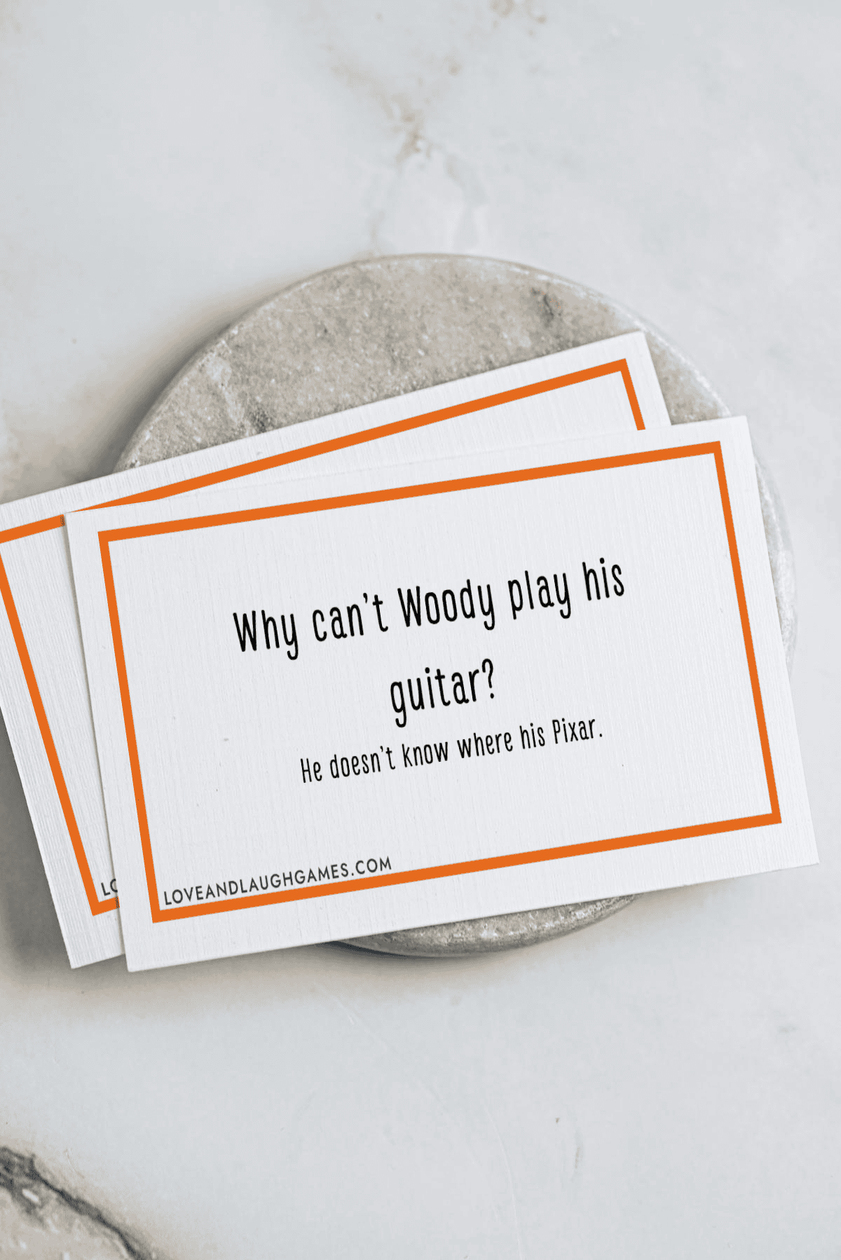 A card reads: “Why can’t Woody play his guitar? He doesn’t know where his Pixar.” The card has an orange border and rests on a round marble surface, with another similar card partially beneath it.