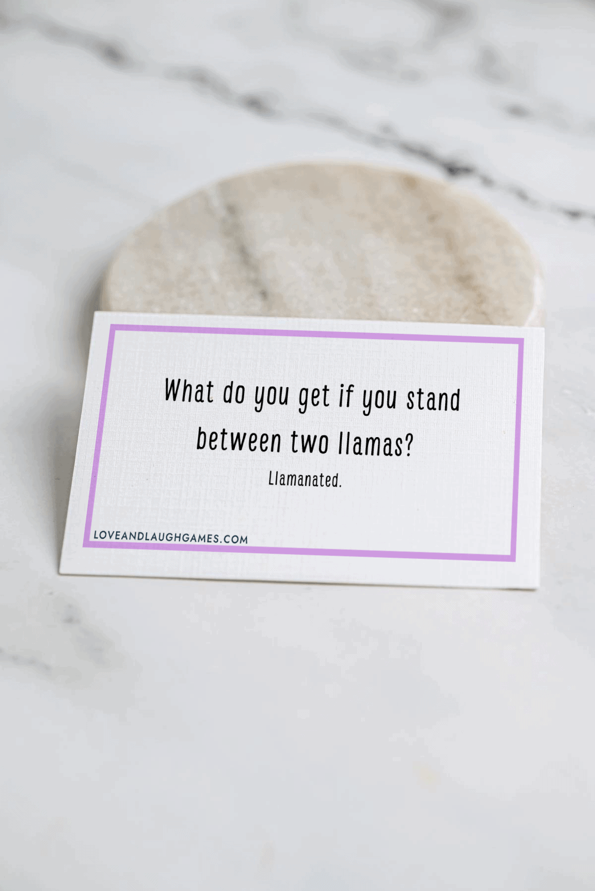 A card on a marble surface reads, What do you get if you stand between two llamas? Llamanated. The card has a purple border and a website link at the bottom: loveandlaughgames.com.