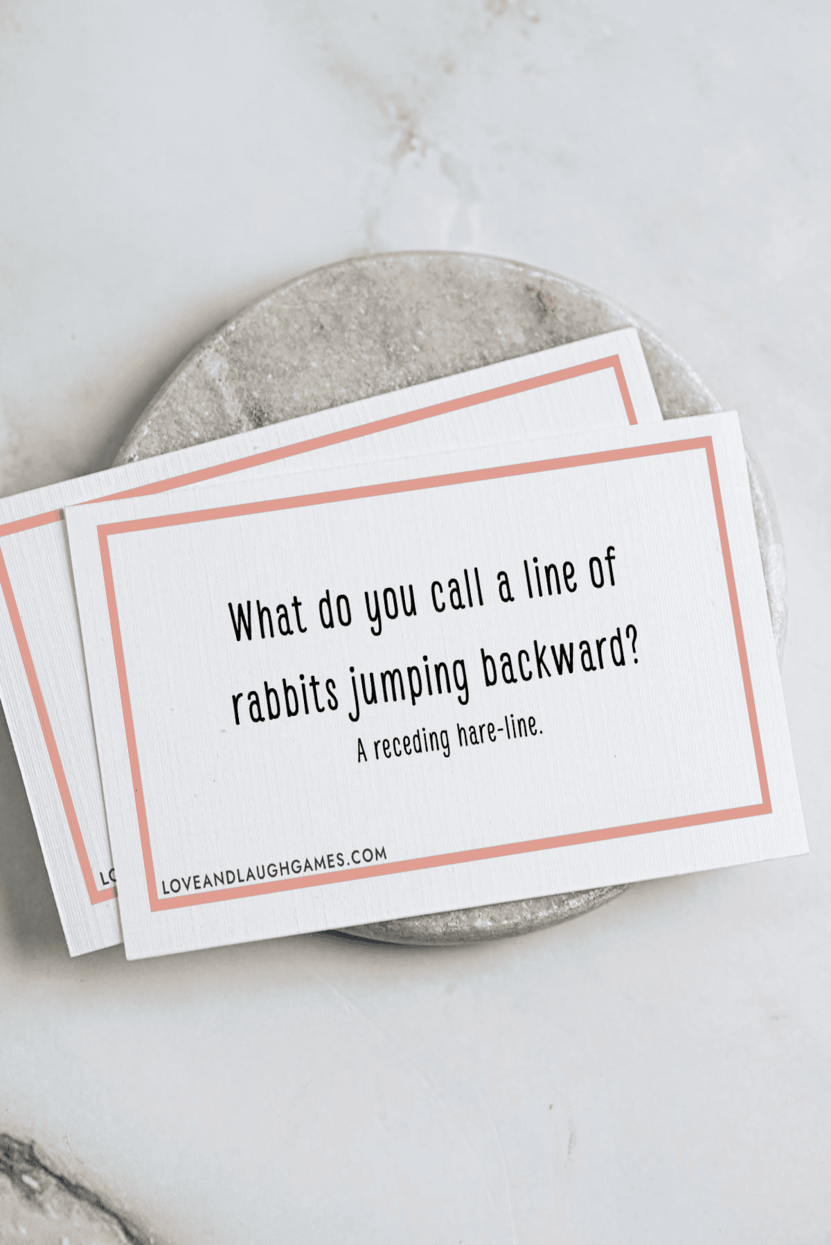 A card on a marble surface reads: “What do you call a line of rabbits jumping backward? A receding hare-line.” Another similar card is partially visible underneath.