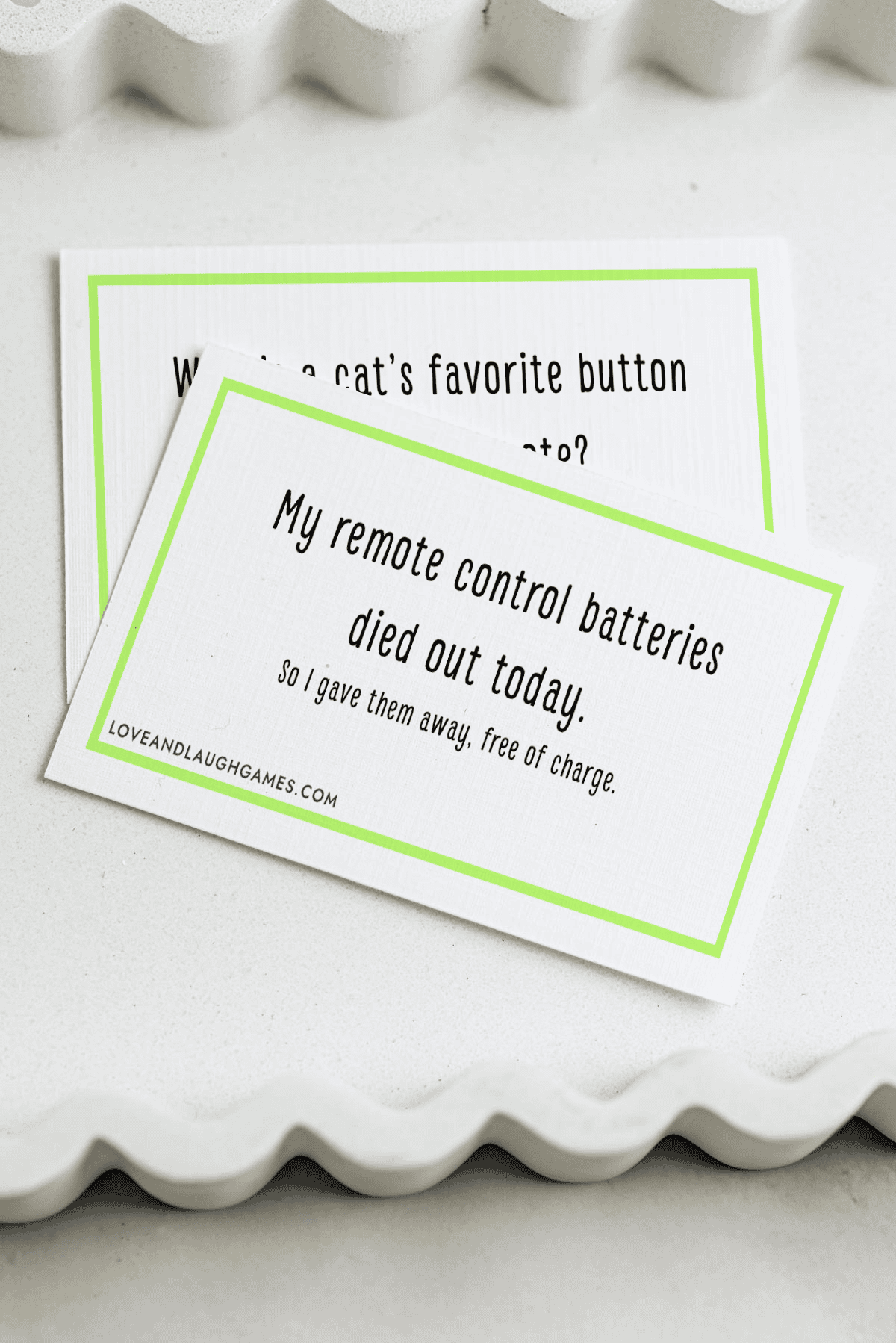 A card with a remote joke reads: My remote control batteries died out today. So I gave them away, free of charge. Another partially hidden card rests beneath it, both displayed on a white, wavy-edged tray.