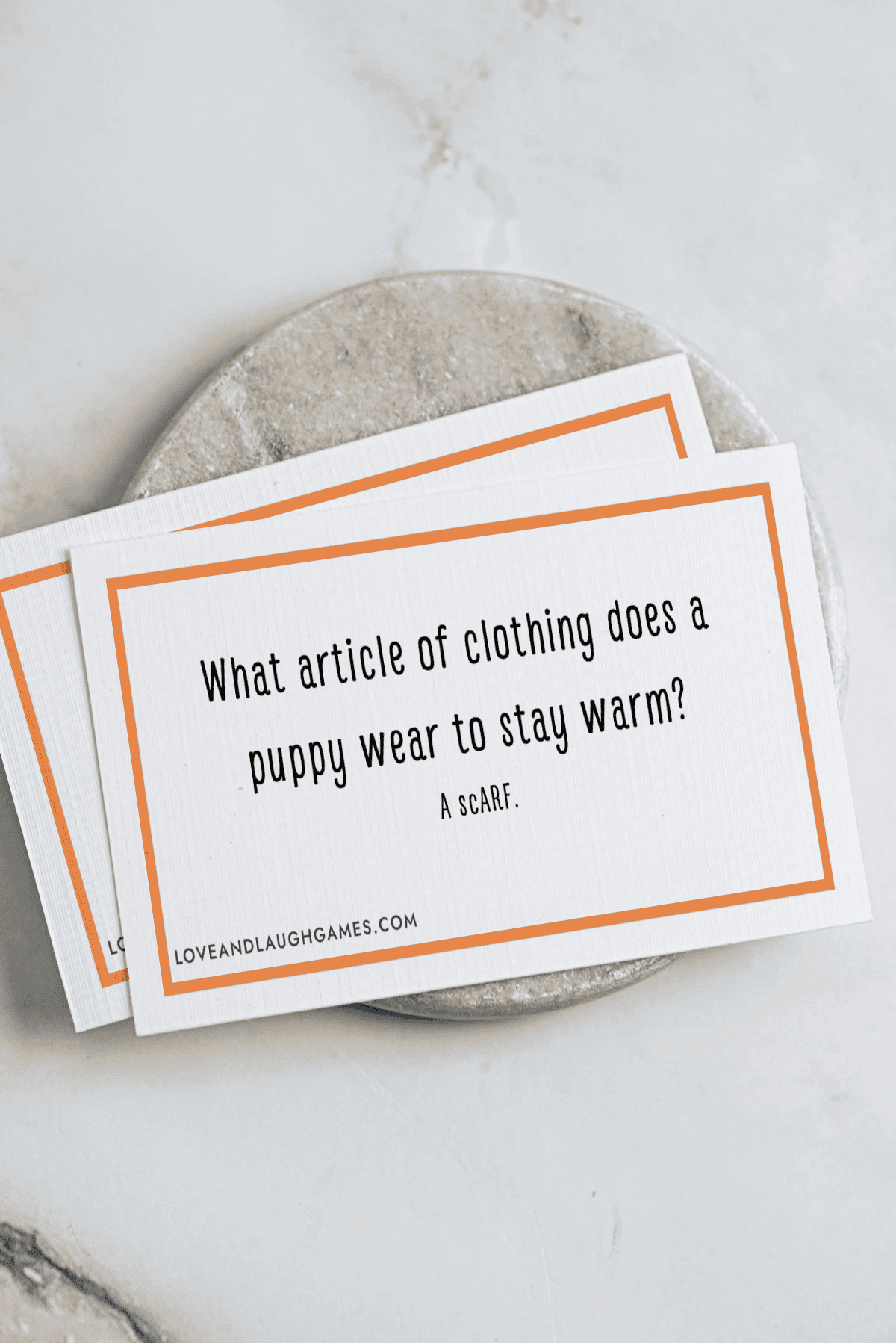 A card on a marble surface reads: What article of clothing does a puppy wear to stay warm? A scarf. The card has a simple border and text layout.