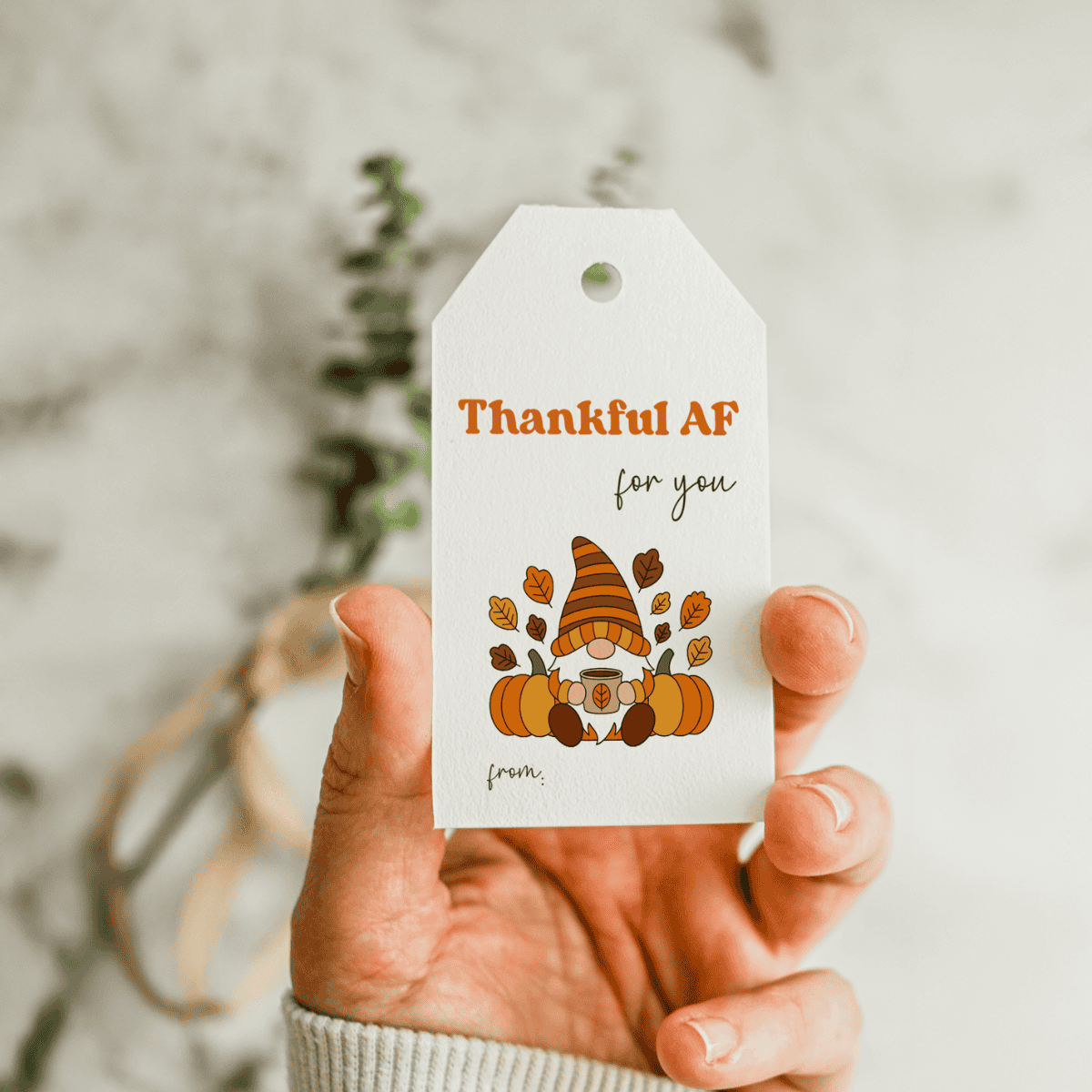 A hand holds a gift tag featuring the text Thankful AF for you above a gnome with orange hat, pumpkins, and autumn leaves. The tag has a from section at the bottom.