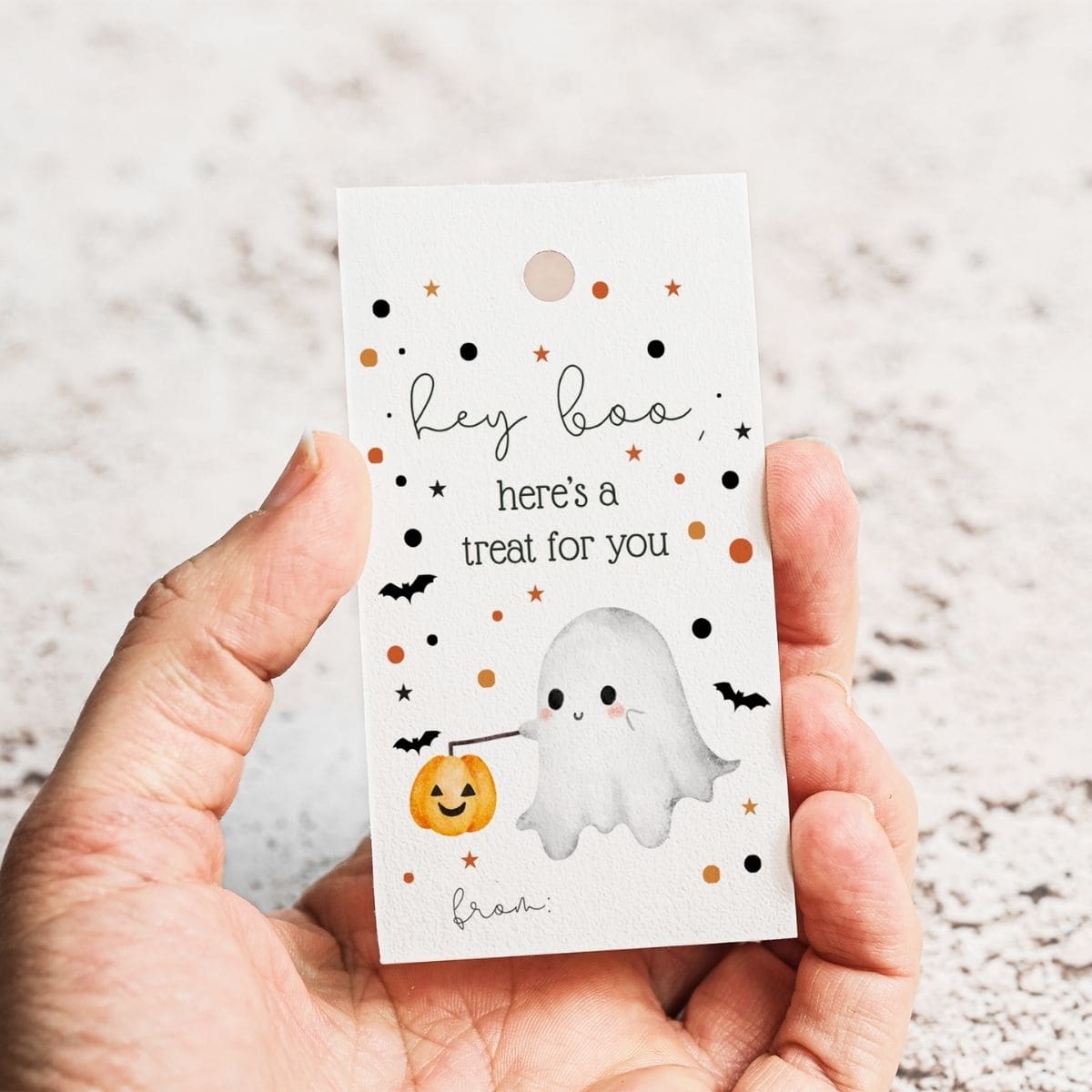 A hand holds a card with cute Halloween art of a smiling ghost holding a pumpkin bucket, surrounded by stars and bats. These Printable Halloween Gift Tags read: hey boo, here’s a treat for you.
