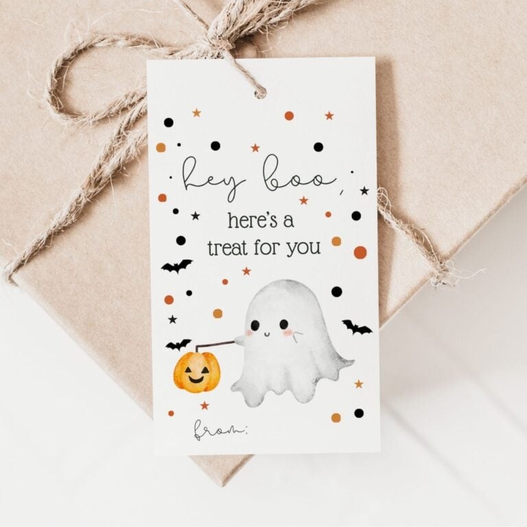 A printable Halloween gift tag featuring a cute ghost holding a pumpkin, surrounded by bats and confetti, reads: Hey boo, here’s a treat for you, with from: at the bottom, attached to a brown paper-wrapped gift with twine.