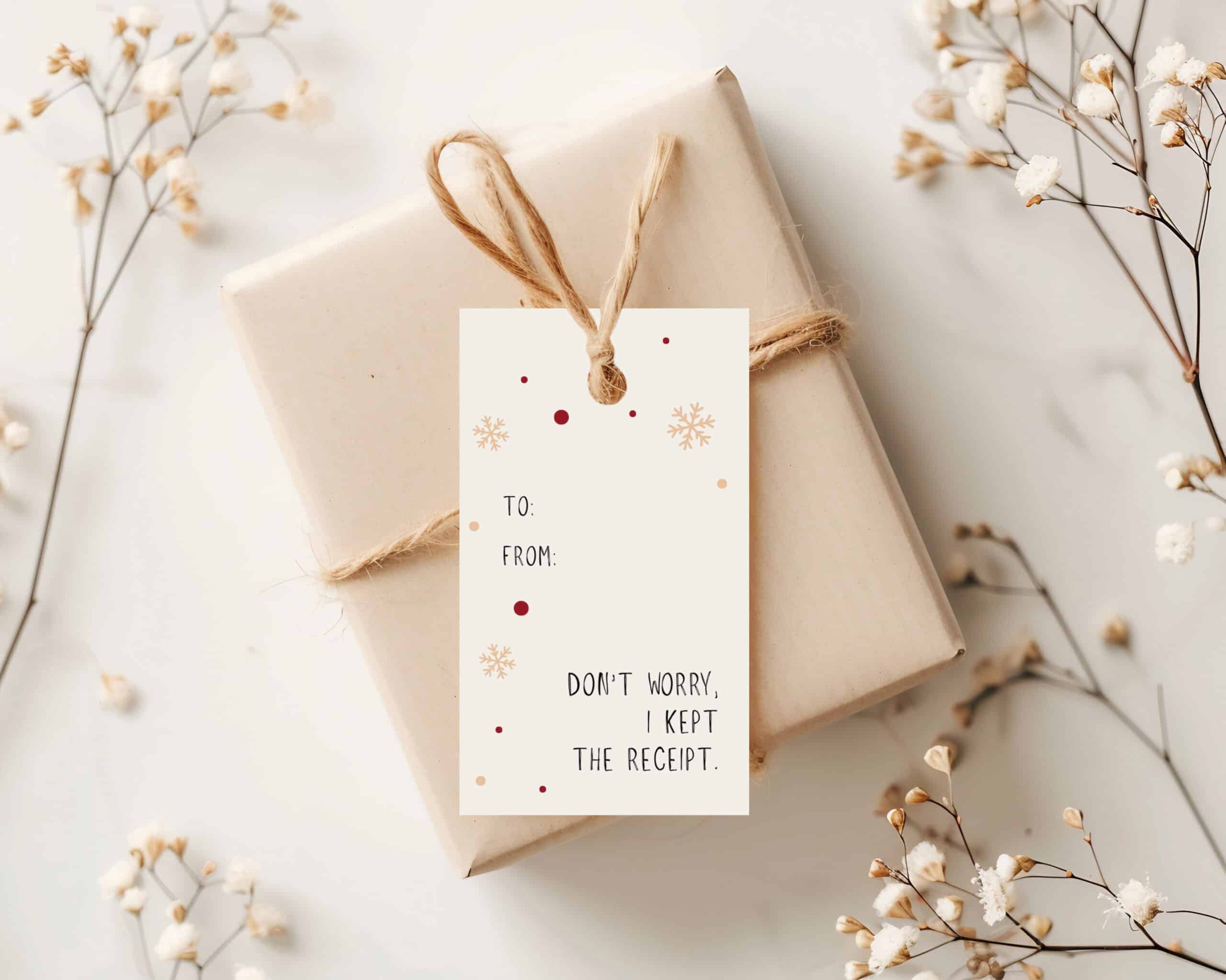 A neatly wrapped gift with twine sits on a white surface, surrounded by small white flowers. A festive gift tag reads, “To: From: Don’t worry, I kept the receipt,” accented with snowflake and dot decorations.