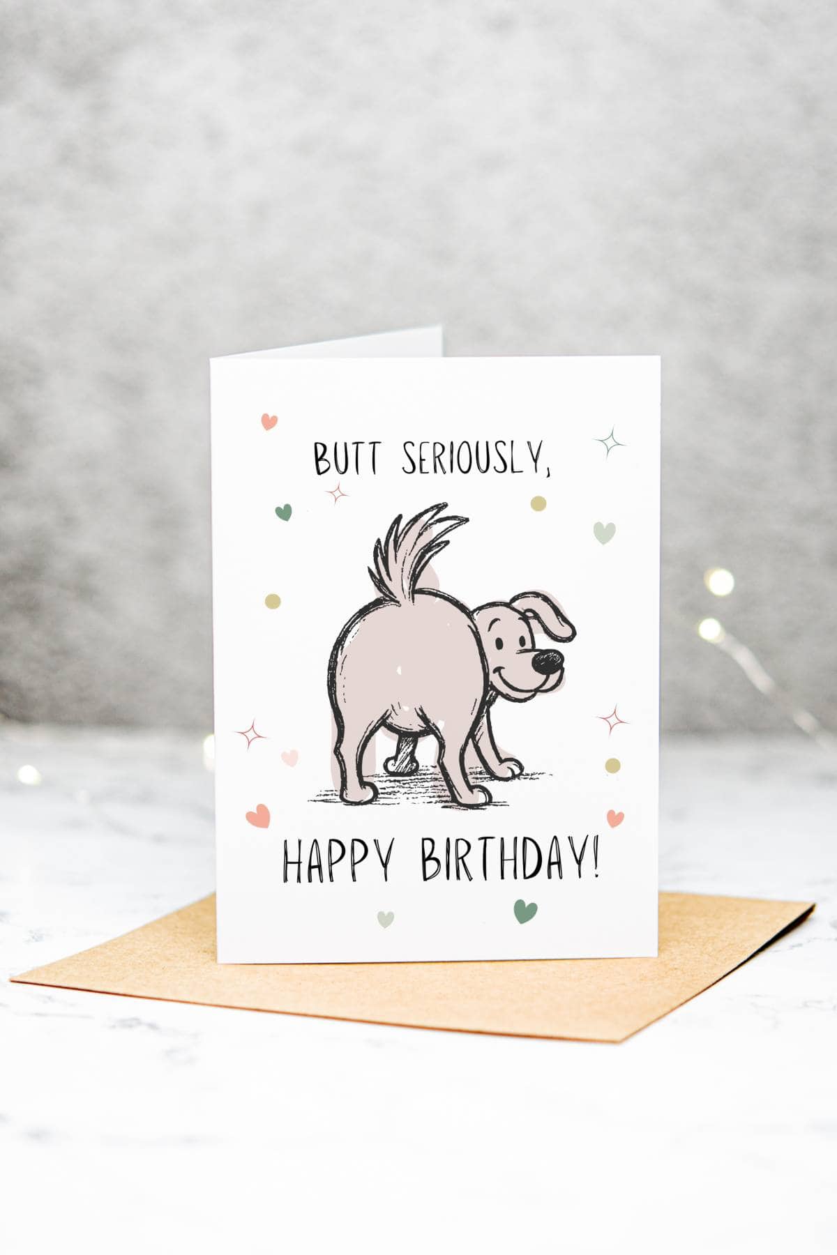 A birthday card featuring a cartoon dog showing its rear with the text BUTT SERIOUSLY, HAPPY BIRTHDAY! surrounded by small colorful hearts and stars, standing on a brown envelope.