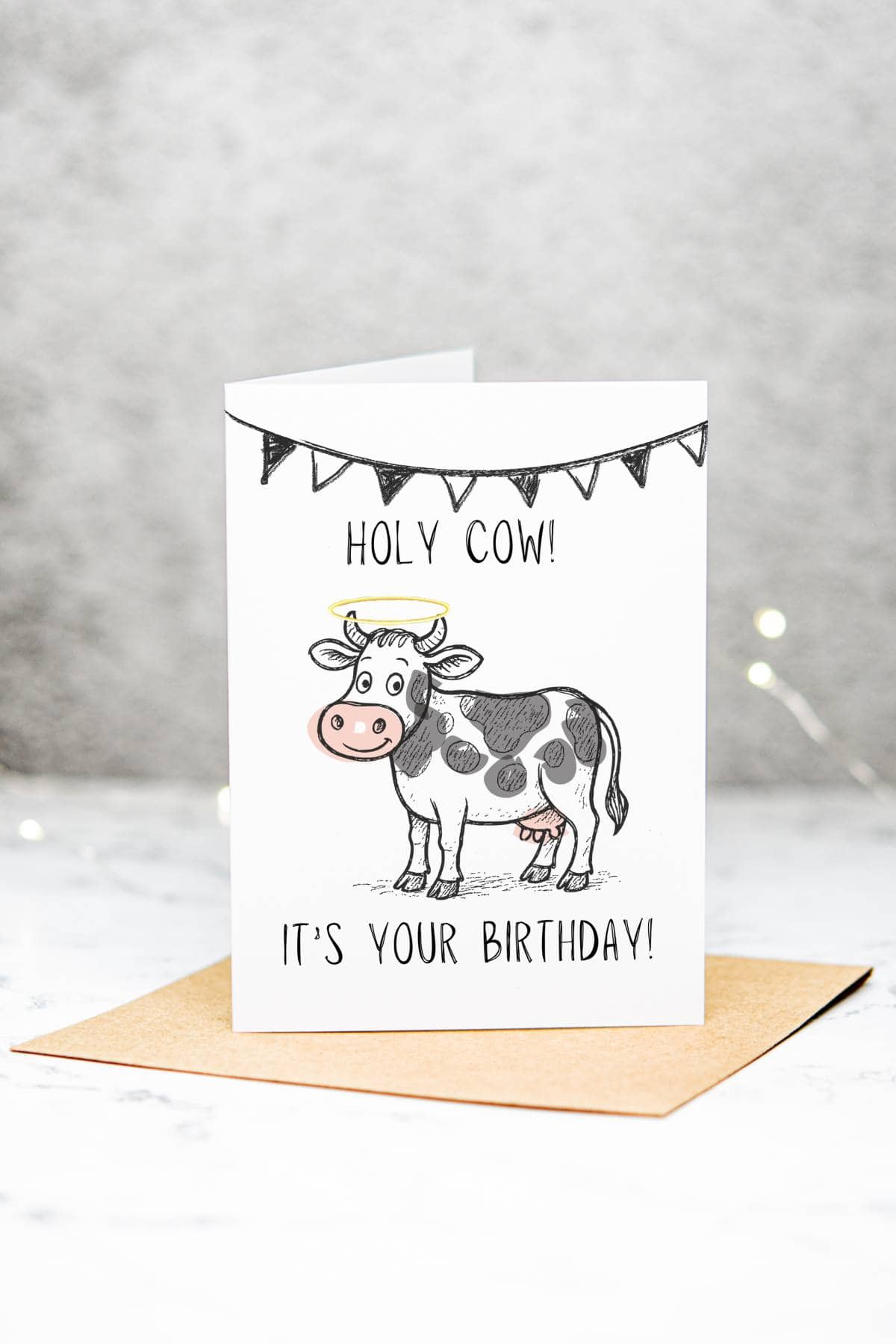 A birthday card featuring a cartoon cow with a banner above it. The card reads “HOLY COW! ITS YOUR BIRTHDAY!” and is standing on a brown envelope against a light, blurred background.