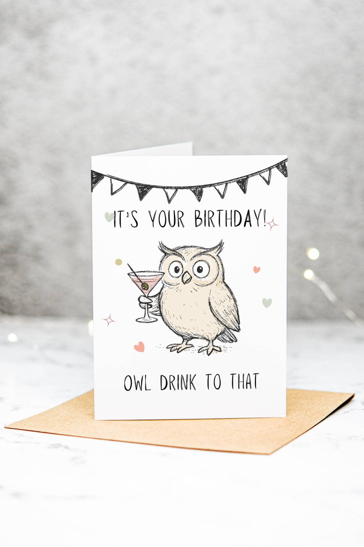 A birthday card featuring a cartoon owl holding a cocktail. The card reads, Its your birthday! Owl drink to that, with small hearts and a black bunting banner above the text. The card is on a brown envelope.