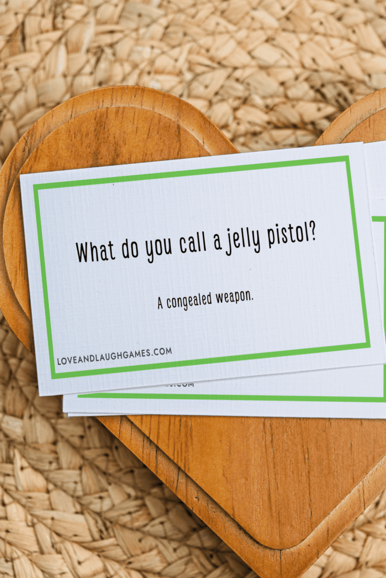 A card reads, What do you call a jelly pistol? A congealed weapon. Part of the delightful Jelly Jokes collection, the card rests on a wooden heart-shaped board atop a woven surface.