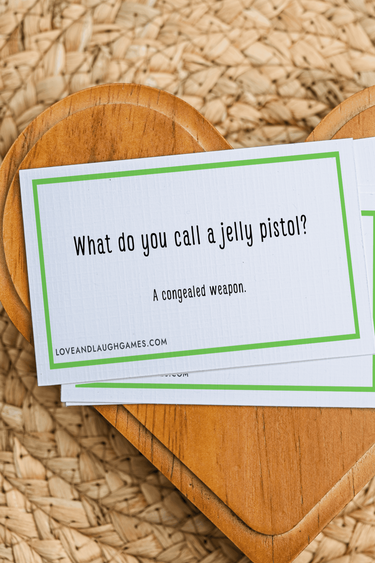 A card reads, What do you call a jelly pistol? A congealed weapon. Part of the delightful Jelly Jokes collection, the card rests on a wooden heart-shaped board atop a woven surface.
